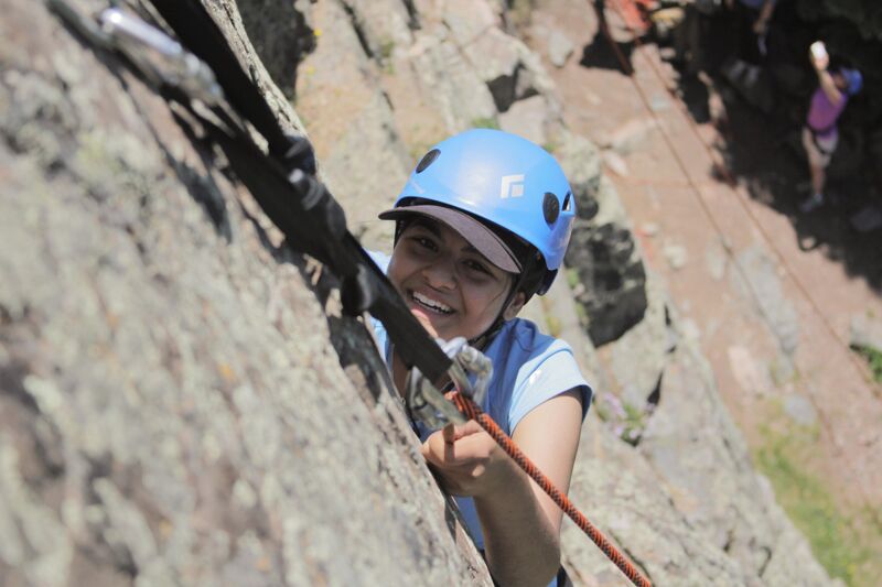 A young woman is rock climbing, wearing a blue helmet and a harness. She is smiling and appears to be enjoying the activity. The rock face is textured and rugged, and there is climbing equipment visible, including ropes and carabiners. In the background, there are hints of greenery and other people, suggesting an outdoor climbing location.